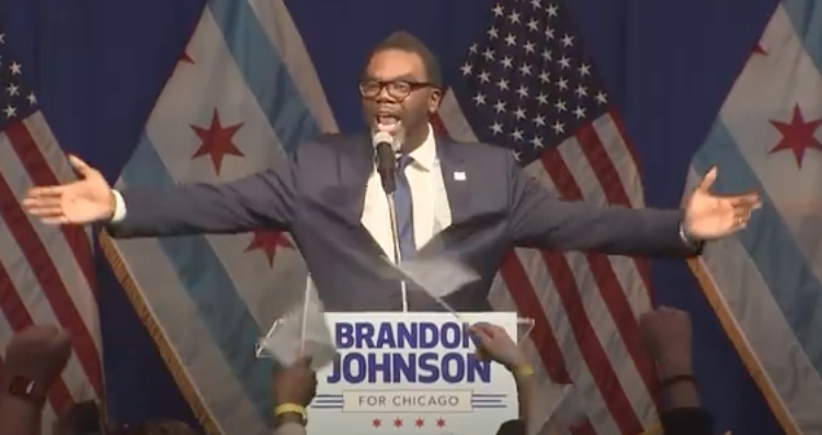 A victorious Brandon Johnson spreads his arms wide as he encourages his supporters to say “…now they know” along with him. --Screenshot from a television newscast